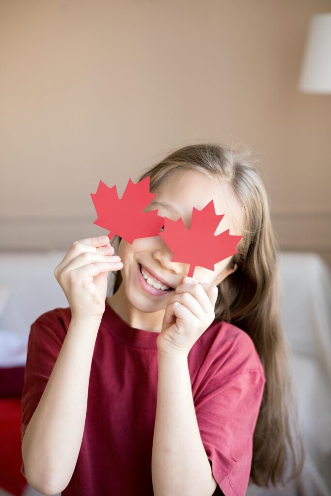 pexels-photo-7144660-7144660 Happy child holding paper maple leaves, celebrating Canada Day indoors.